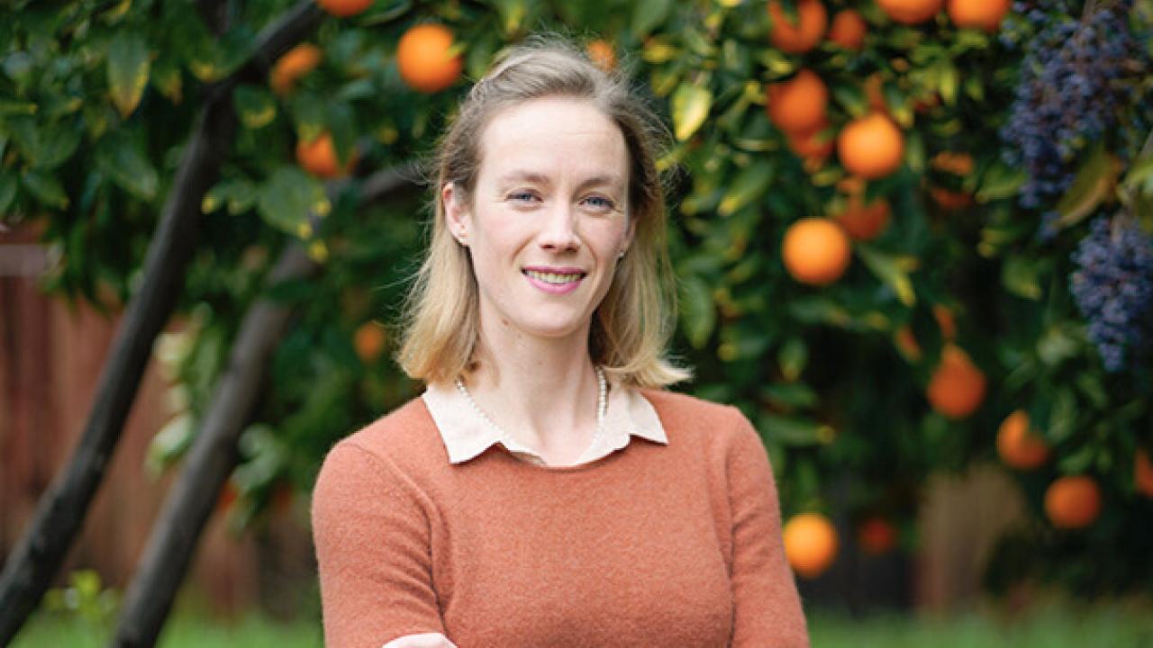 Paige Kouba stands in front of an orange tree. She is wearing an orange sweater over a light colored button up shirt. 