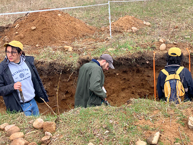 Three young people working in a long, deep trench in tan-colored soil.