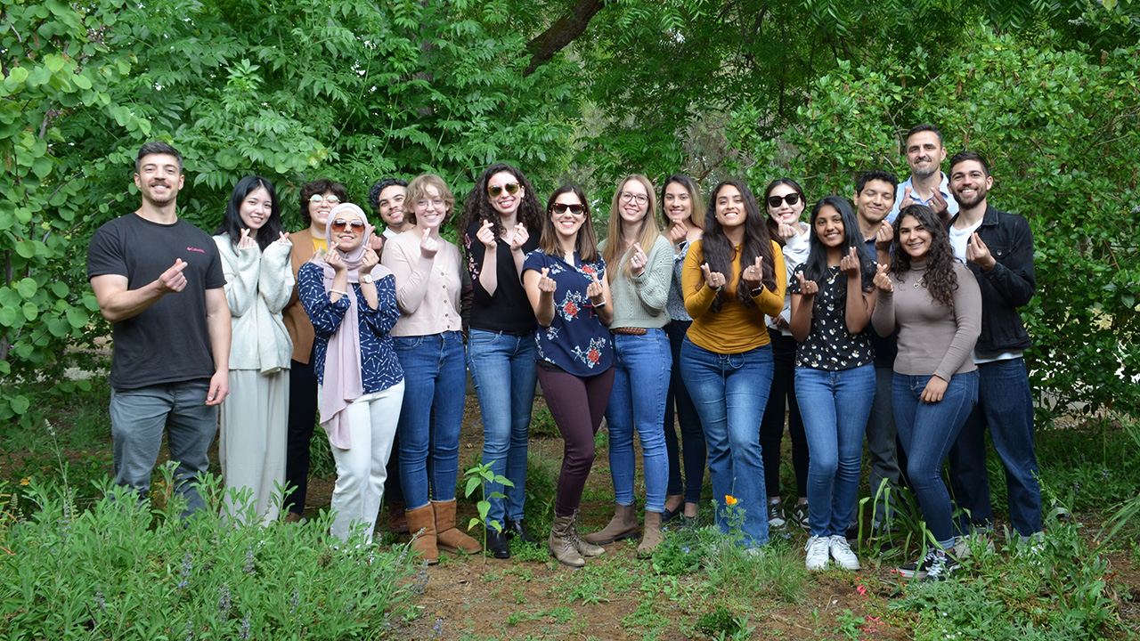 17 people in a line, outdoors by tall leafy green bushes. Each is smiling and has their thumb and forefinger making a heart shape.