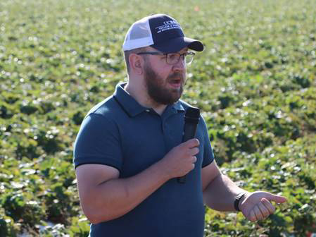 Man wearing a ballcap & holding a microphone while standing in a field of low green plants in furrows