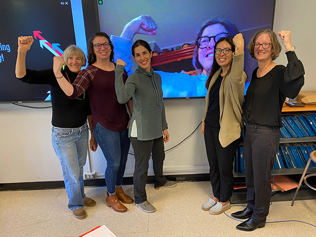 Five women in a room, and a sixth appearing on a large TV screen behind them. They are smiling and holding up clenched fists in a "We are strong!" pose.