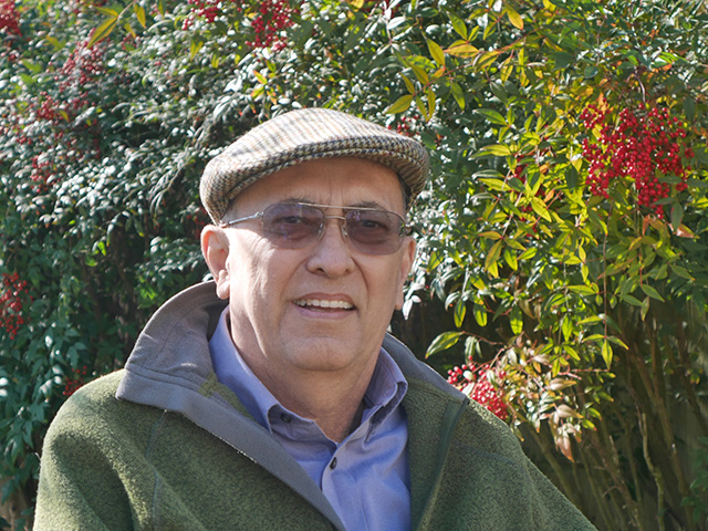 Close-up shot of a man wearing a cap. Behind him are bushes with red berries.