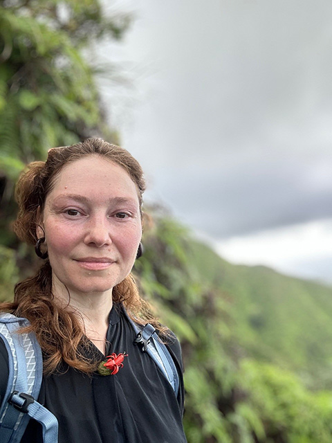 close up of a young woman. Behind her is a steep, vegetation-covered slope and hazy ocean beyond, grey cloudy skiy above.