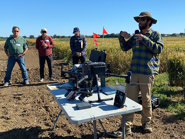 A man talking out in a field of grain. A table in front of him has a four-legged flying device on it and other equipment.