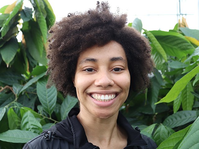 Close-up of a woman smiling, with large green leafy plants in the background.