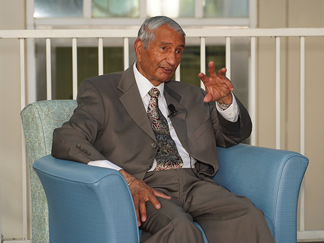 Older man wearig a suit and sitting in a chair, gesturing with his left hand.