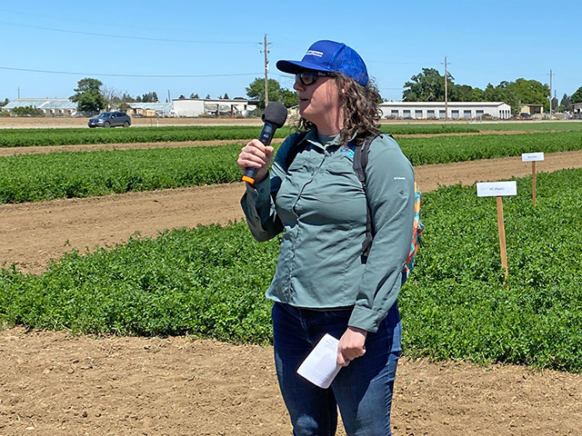 Woman standing in front of a field of low green bushy plants, with a microphone in her hand. Blue sky above