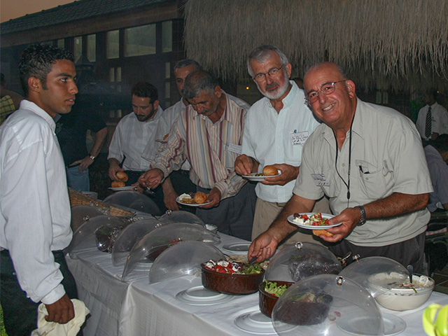 Men standing in front of tables with bowls of food, serving up their plates.