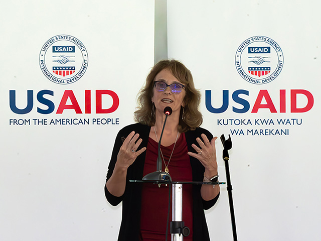 A woman speaking. Behind her, a panel reads "USAID" in English and an African language.