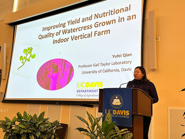 A woman speaking at a podium, with a slide projected on a screen behind her that reads, "Improving Yi8eld and nutritional quality of watercress grown in an indoor vertical farm.'