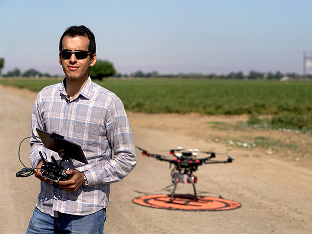 Man holding a control device in his hands, with a drone behind him on an organge landing circle on a dirt strip between green fields