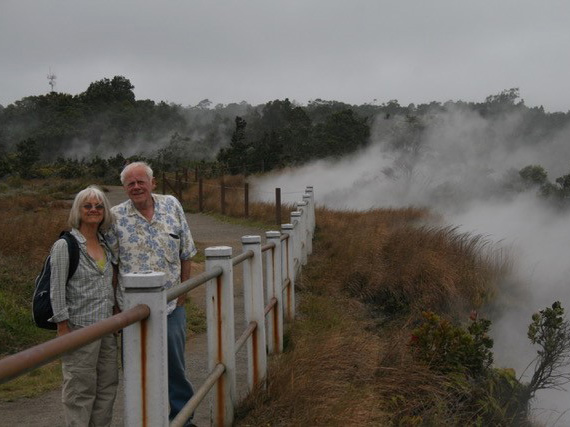 An older man and woman in an outdoor scene, standing behind a short white fence. Beyond the fence, we see the land drops off. Thick, white vapor billows up and away fromo the. Trees in the distance.