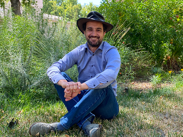 A man wearing a hat and sitting cross-legged on grass in a tree-filled courtyard