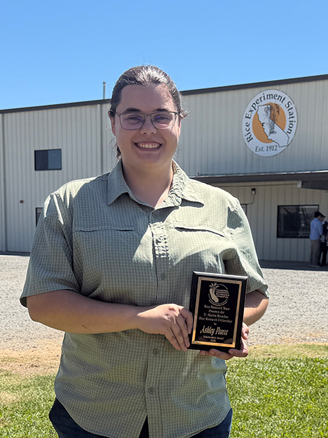 Smiling young woman holding award plaque outside a large metal building with round logo that says, "Rice Experiment Station, Established 1912."