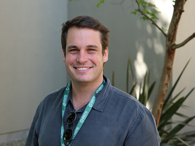 Smiling man in a dark shirt with sunglasses on lanyard, standing outdoors by plants
