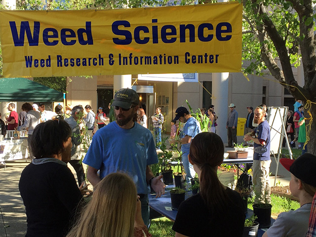 A man outdoors under a tent canopy, talking to people. Above, a yellow sign reads, "Weed Science."
