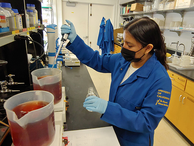 A young woman seated in a lab and wearing a blue lab coat reaches for some liquid