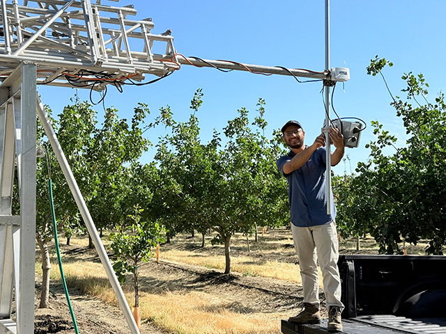 Man in an orchard. He's holding a white metal pole that is part of a larger contraption of white metal bars built into a tower-like structure, quite a bit taller than he is.