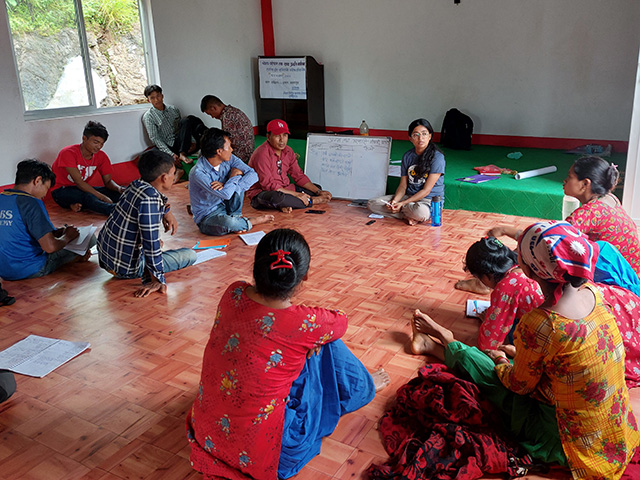 12 men and women sitting on the floor in a room, the men on one side and the women on the other. The women are wearing bright and colorful clothing.