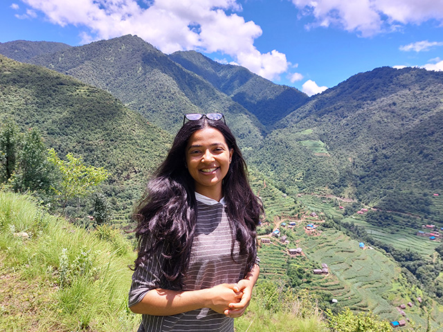 A young woman standing, with high mountains and a cloud-studded blue sky in the background.