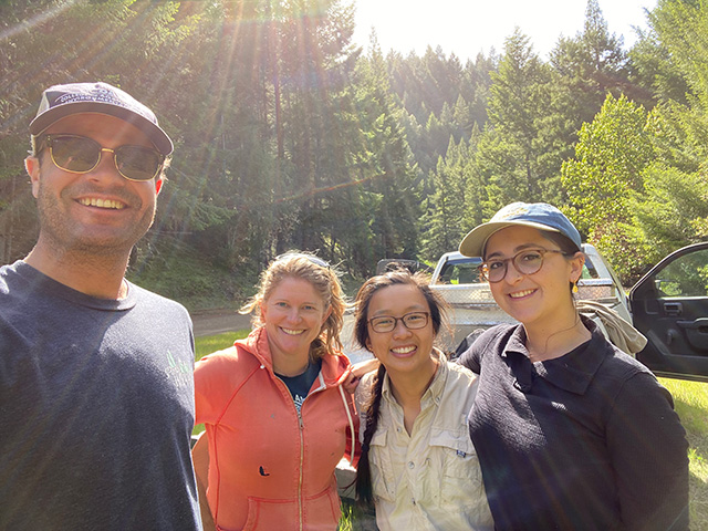 Four smiling adults standing in a sunlit grassy clearing with forest behind