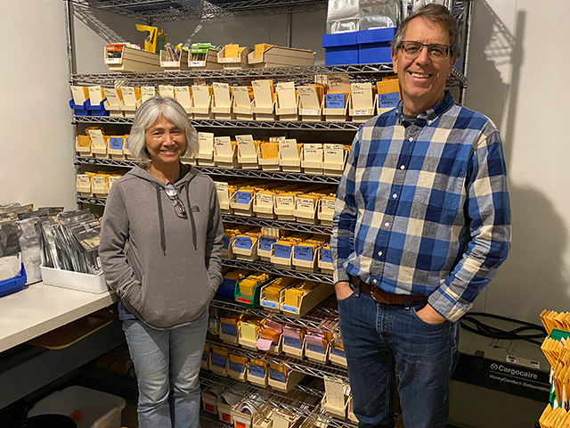 A woman and a man standing in a small room. Behind them is a rack with many shelves. Small brown paper packets sit in boxes on the shelves.