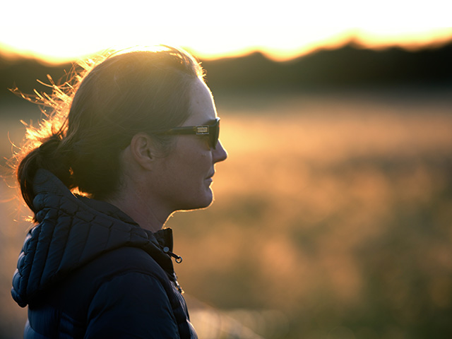 Close-up of woman looking into the distance. Orange sunlight reflects off the grassland behind her.