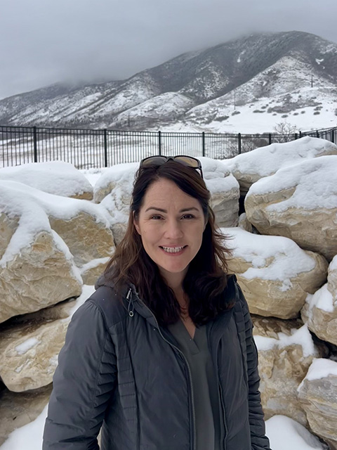 Woman in foreground by a snow-covered, low stone wall. Behind her is a snow-covered field, and at the near horizon is a snowy mountain with gray clouds hanging near.