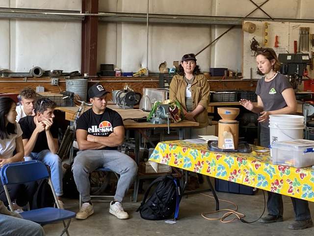 A young woman stands at a table with a small mill, while seated students look on, all inside a metal building.