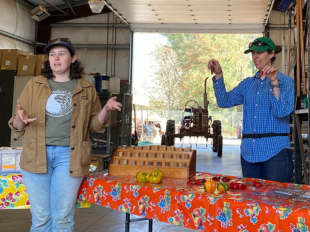 Two young women stand at a table, one of them holding up green jalapeno peppers, and the other one talking.