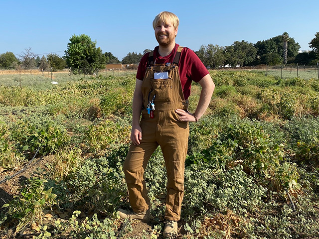 A young man wearing overalls stands in a field, against a clear blue sky.