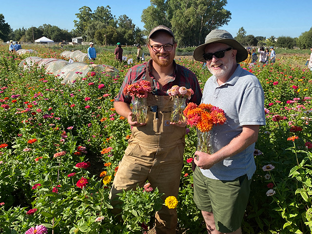 Two men standing in a field of flowers, holding vases with cut flowers.