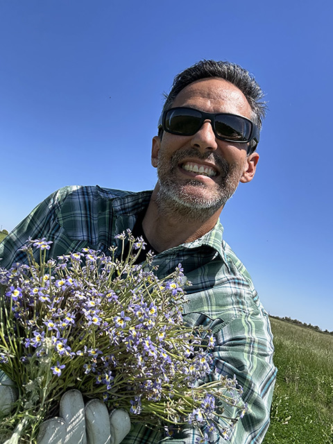 A young man holds a purple flowering plant. Blue sky above.