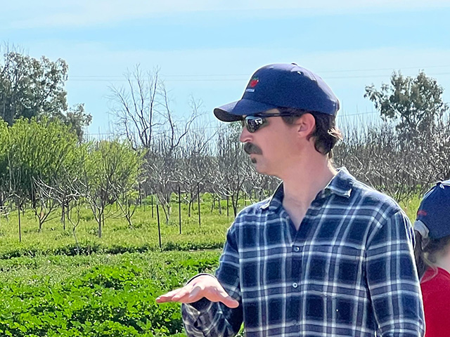 Man in an orchard with green grassy plants between rows of trees. He's gesturing with his hand.