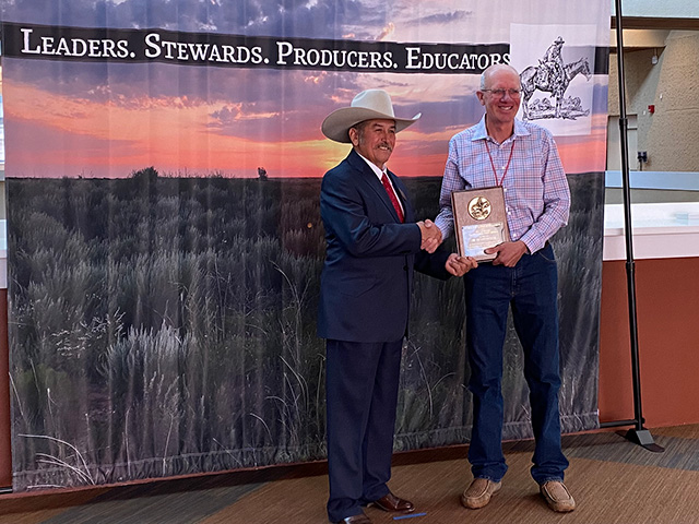 Man in a cowboy hat smiles as he gives an award to another man and shakes his hand. Sign behind them says, "Leaders, Stewards, Producers, Educators"