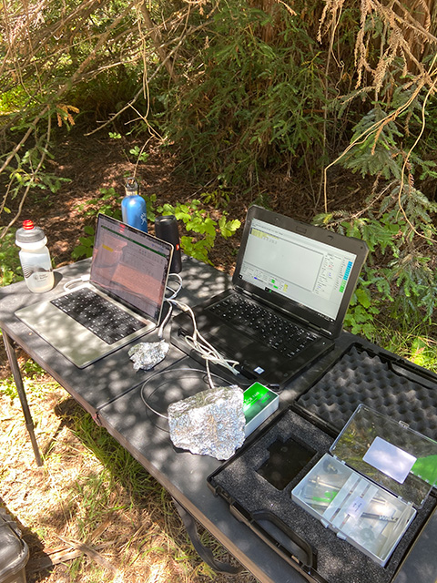 Outdoor field workstation on bench with two open laptops, cables and bottles set up under trees. UC Davis Department of Plant Sciences