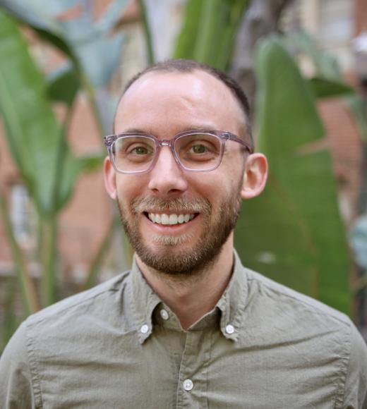 Man in glasses and green shirt smiles for camera. Green leaves in background.