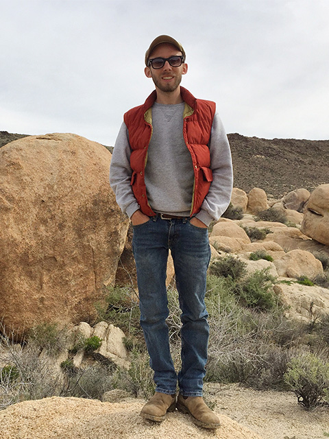 A man dressed for hiking, standing on a rock in a landscape of dry grass