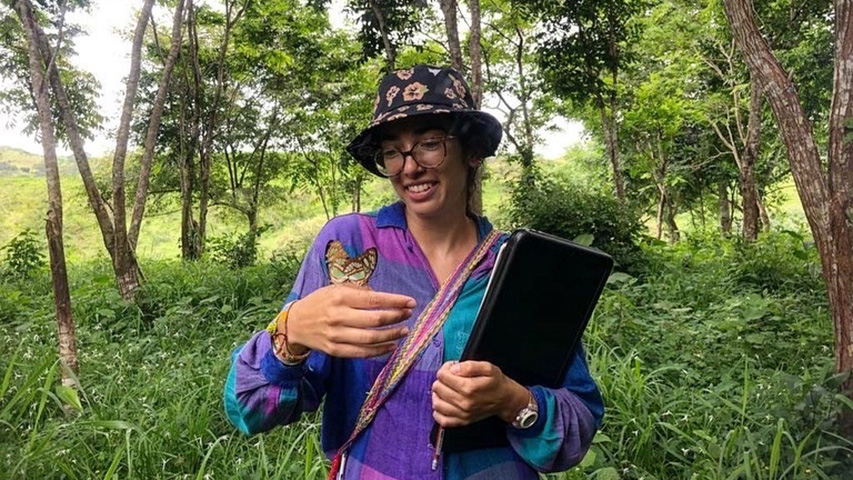 A young woman wearing colorful lavender-and-turqoise clothing stands in a small clearing surrounded by tropical trees. A brightly colored butterfly sits on her hand, as she looks at it and smiles.