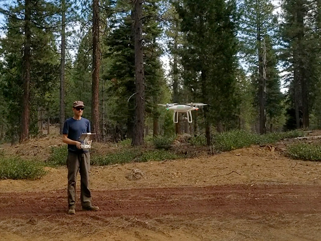 A man holds a remote controller while a drone flies higher than his head, with forest trees in the background.