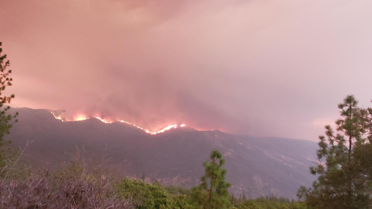 Orange-gray smoke billows above green trees, as flames rise from a mountain ridge.