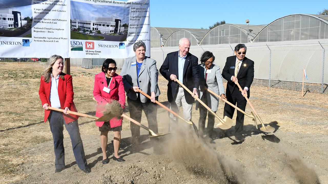People in business attire standing in a field holding shovels and flinging dirt into the air. Behind them are greenhouses behind a fence.
