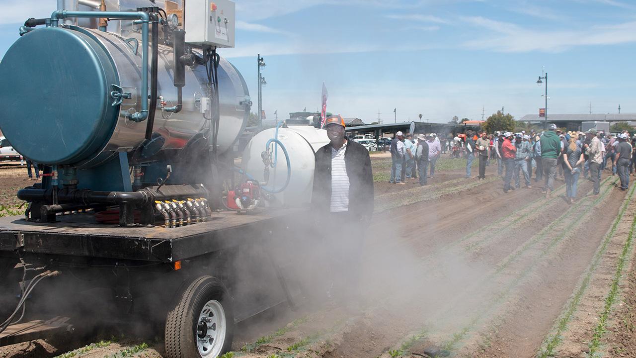 A large, tractor-like machine kicks up a cloud of whitish vapor in a field, while a man stands next to it smiling.
