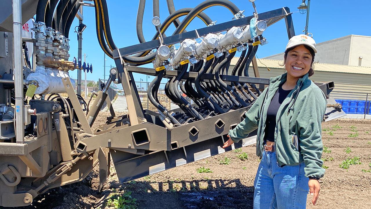 Young women standing by a large tractor-like contraption with an uplifted metal arm, and tubes and things are hanging down from the arm.
