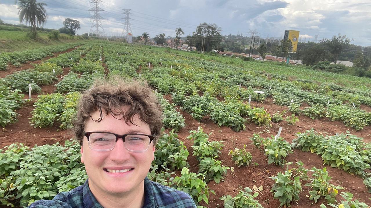 Selfie of a young man. Behind him, a field of green busy plants stretched out to trees, out-buildings and high-voltage towers in the distance.