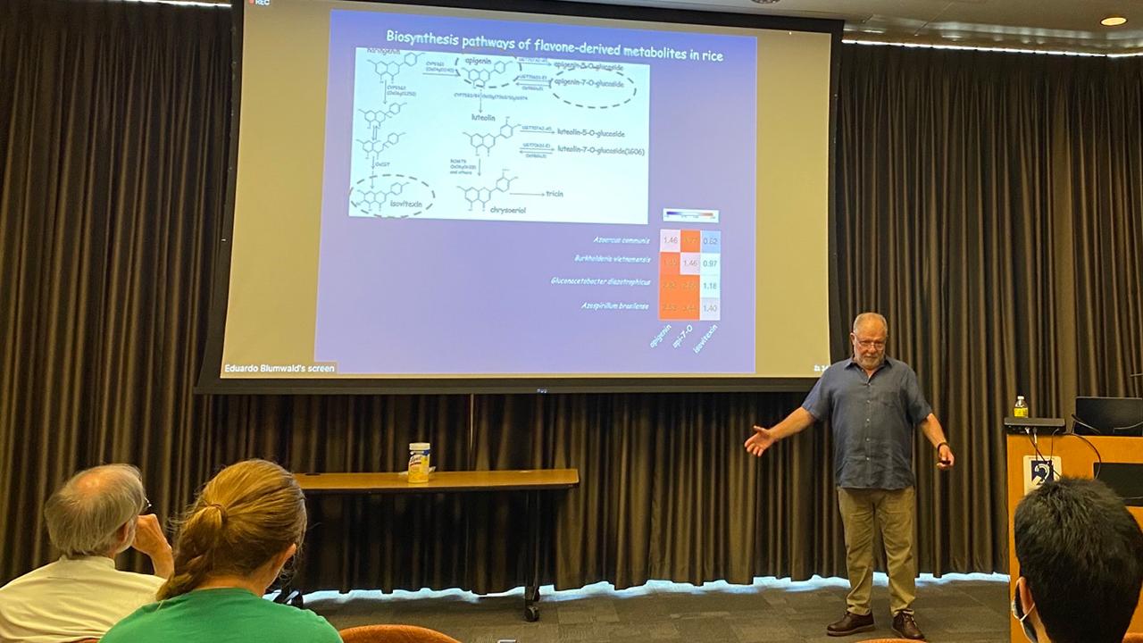 A man gestures in front of a screen with a projected image. People in the audience observe.