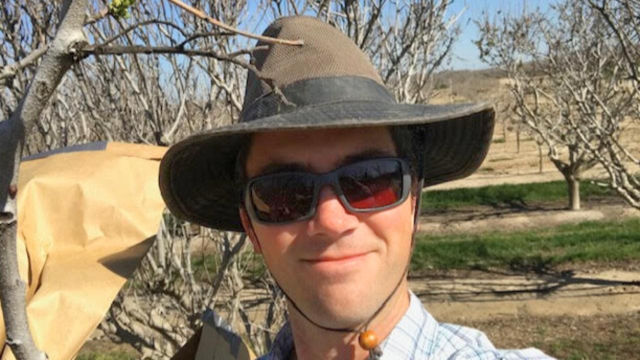 Brown smiles at the camera. He is wearing a brown hat and white-blue striped button up shirt. There are many trees behind him.