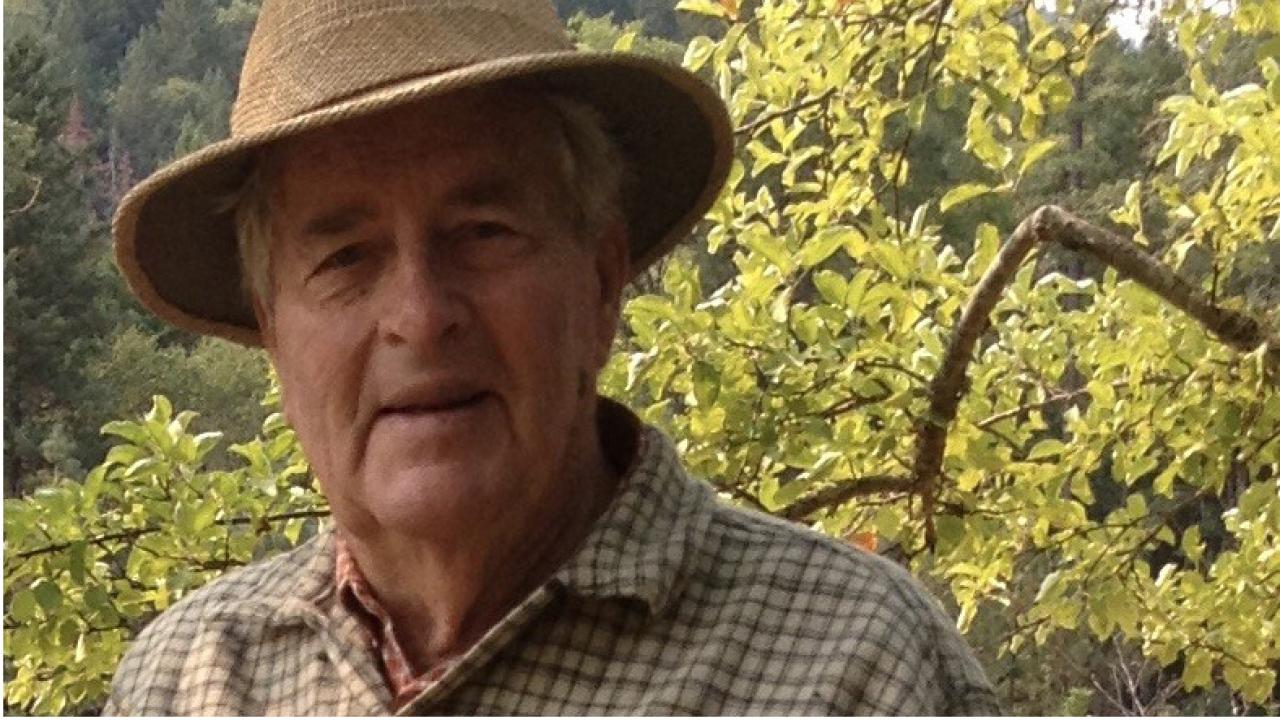 Older man wearing a hat, with a mountainside and trees in the background.