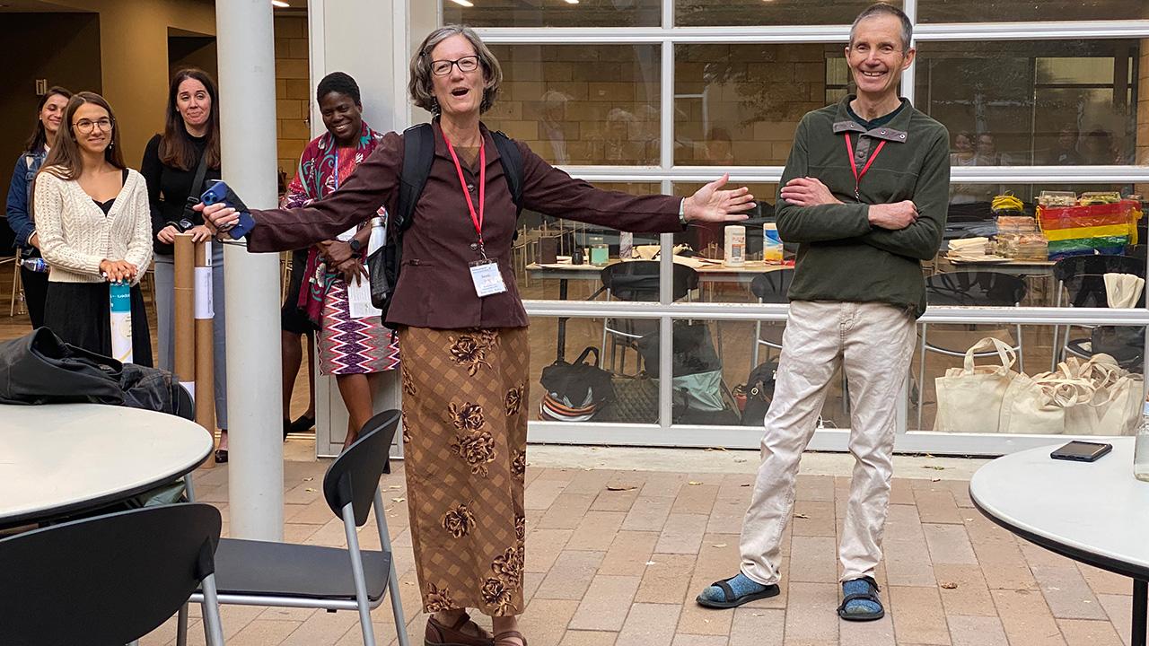 A woman looks joyful, standing with her arms outstretched and smiling in an outdoor patio, speaking to people present.