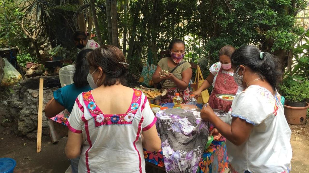 Women wearing traditional embroidered blouses are gathered around an outdoor cooking hearth.